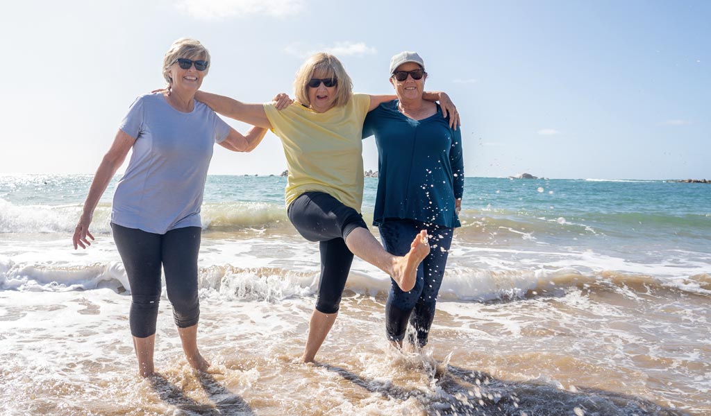 Drei Frauen genießen einen sonnigen Tag, stehen in den Wellen am Strand, lächeln und haben Spaß.