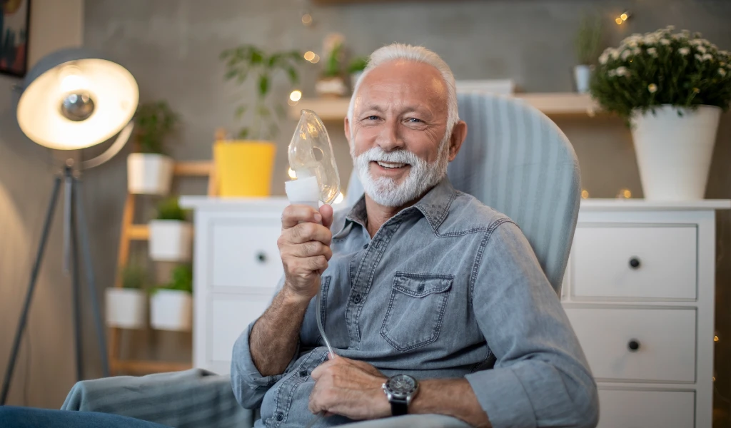 Ein älterer Mann verwendet einen Inhalator zu Hause und lächelt in die Kamera.