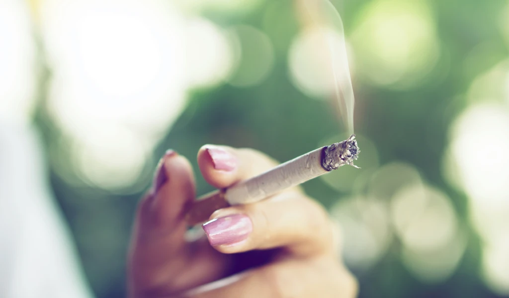 Hand mit rosa Nägeln, die eine brennende Zigarette vor einem verschwommenen grünen Hintergrund hält.