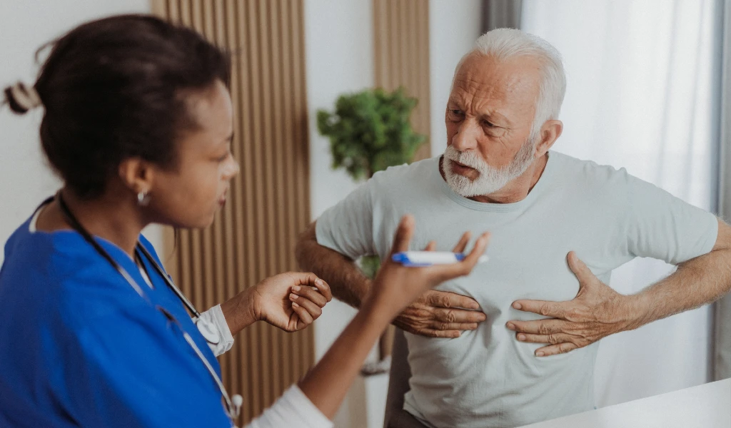 Ein älterer Mann hält sich die Brust und spricht mit einer Ärztin über seine Atemprobleme.