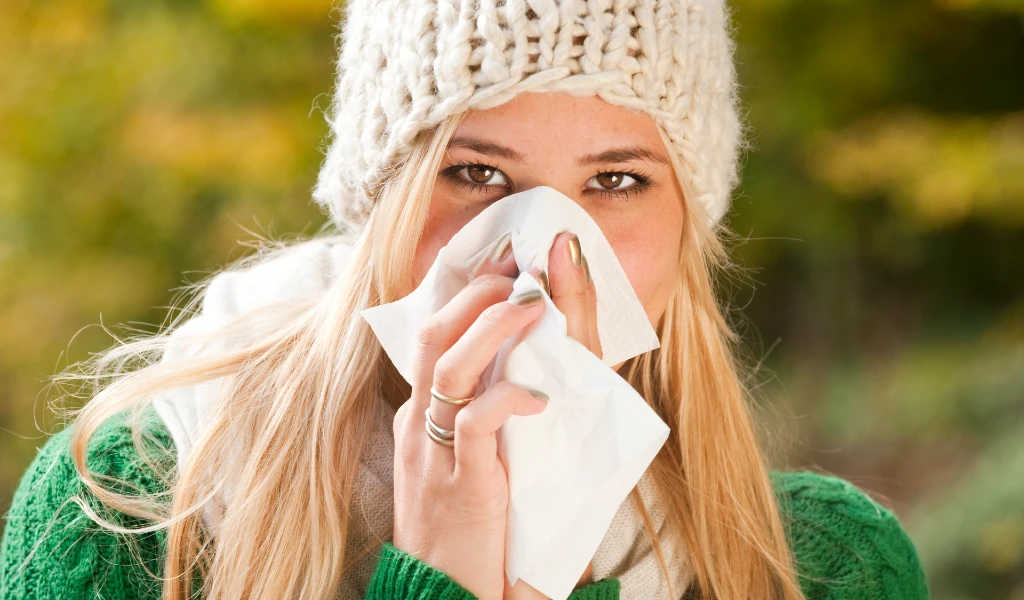 Frau mit Mütze niest in ein Taschentuch, umgeben von blühendem Raps.