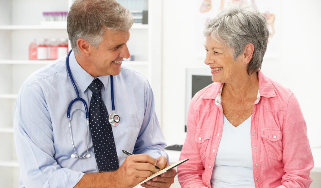Lächelnder Arzt mit Stethoskop notiert etwas, während er einer älteren Frau in rosa Bluse zuhört. Praxisumgebung.
