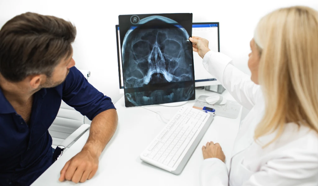 Ärztin zeigt auf Schädel-Röntgenbild am Bildschirm, Patient schaut zu.