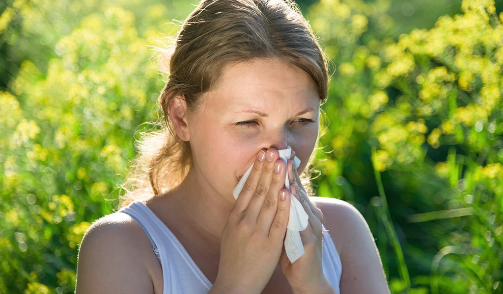 Junge Frau mit Heuschnupfen schnäuzt sich mit einem Taschentuch in einer blühenden Wiese
