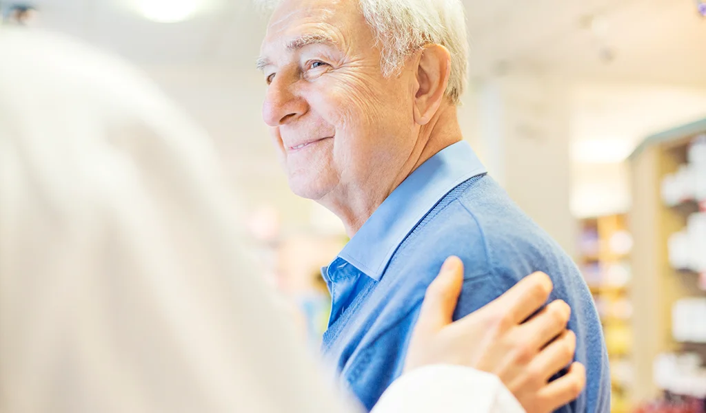 Älterer Mann in einer Apotheke lächelt, während eine Person in weißem Kittel ihm eine Hand auf die Schulter legt.