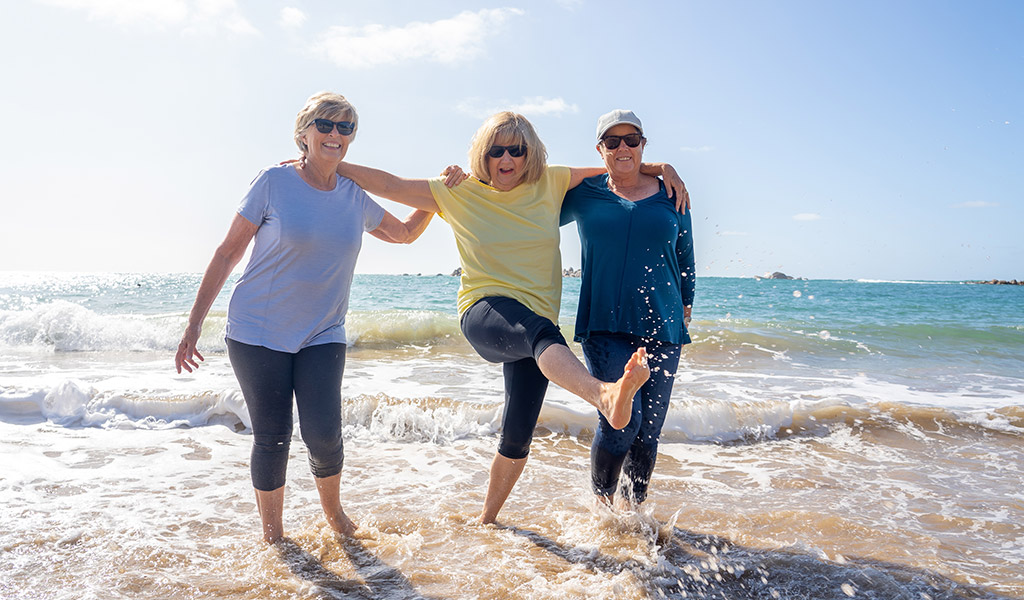 Drei Frauen genießen einen sonnigen Tag, stehen in den Wellen am Strand, lächeln und haben Spaß.
