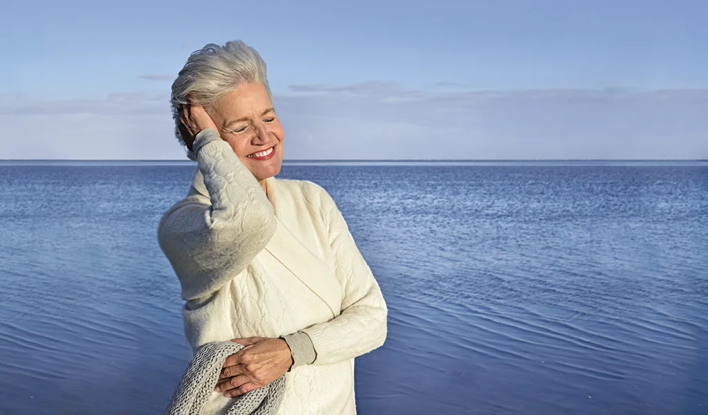 Ältere Frau mit grauen Haaren steht lächelnd am Meer, trägt einen cremefarbenen Pullover und legt eine Hand an ihr Gesicht. Der Himmel ist blau und das Wasser ruhig.